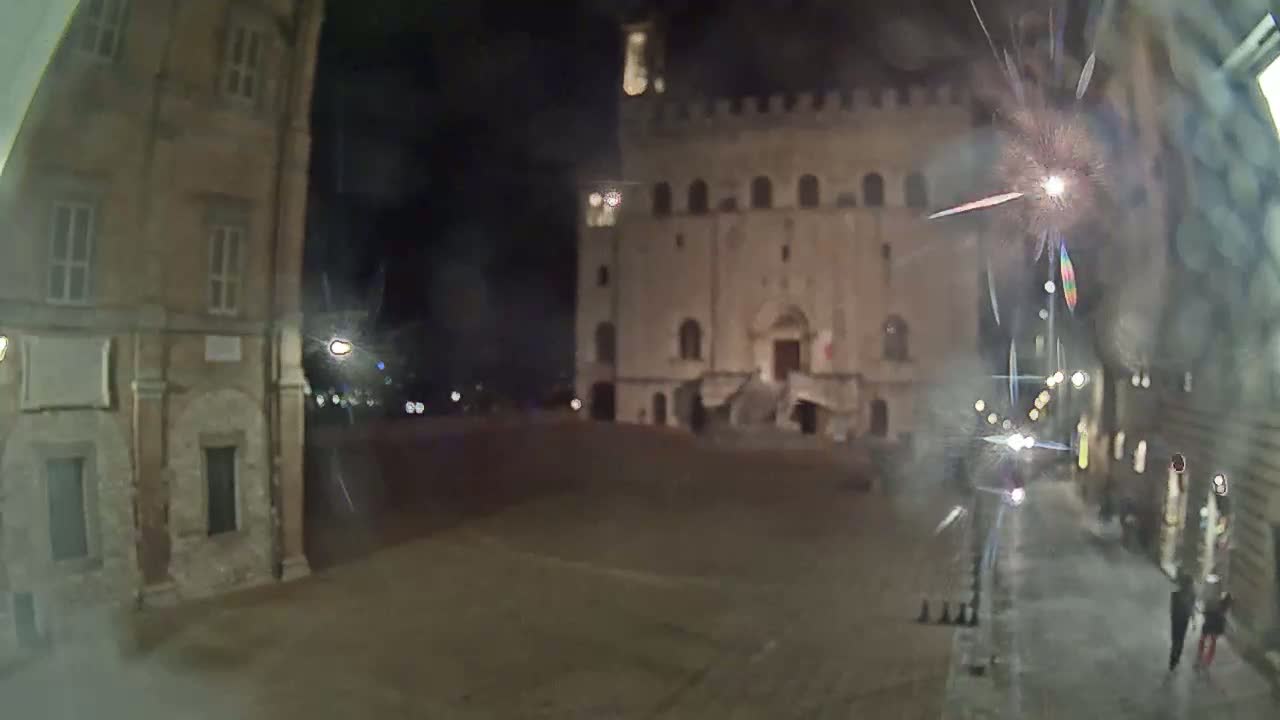 A nighttime, low-angle view of a town square with two large stone buildings, one of which has a large staircase leading to its entrance; fireworks are visible in the sky.