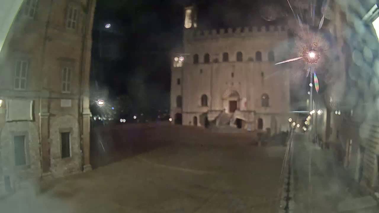 A nighttime view of a town square, showing a large, illuminated building with steps, flanked by older buildings, seen through a cracked window.
