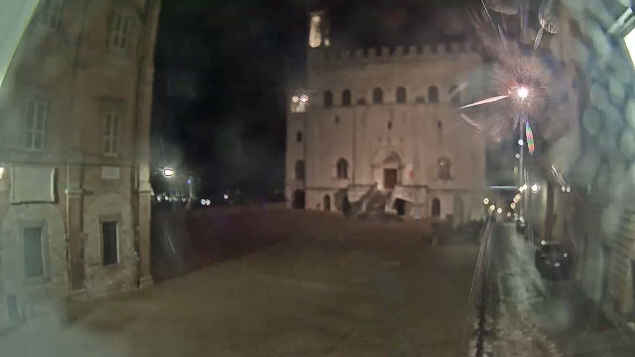 A nighttime view of a town square, featuring a large, illuminated stone building and surrounding older structures, with fireworks visible in the distance.