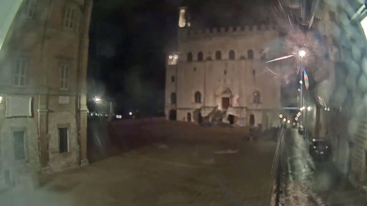 A nighttime view of a large, illuminated building in a square, seen through rain-streaked glass.