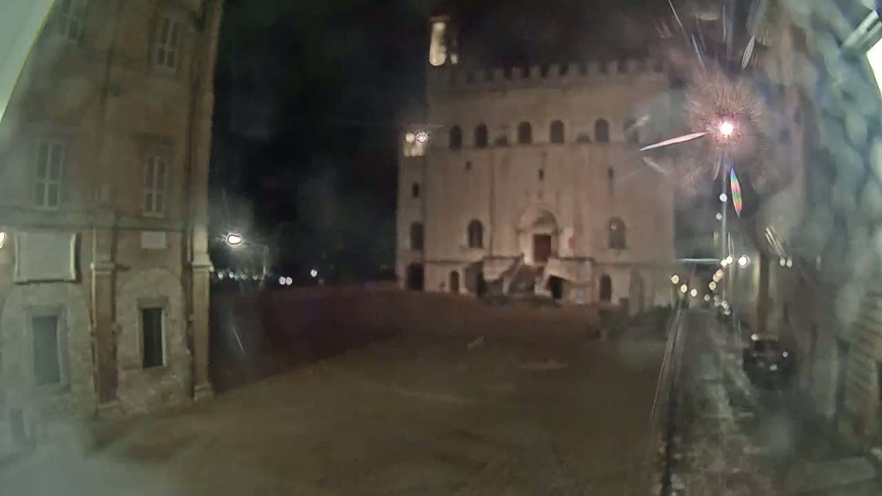 A nighttime view of a town square, showing old stone buildings, one of which is large and has stairs leading to its entrance, under a dark sky.