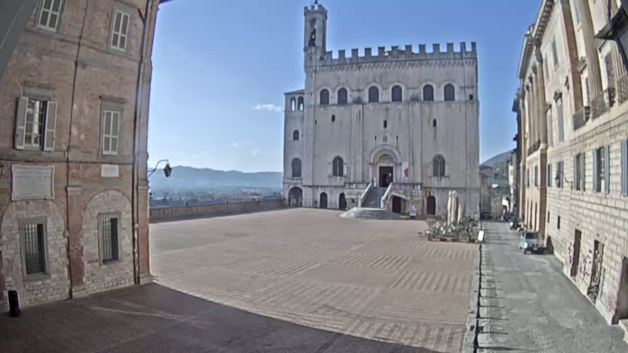 A large, empty plaza is situated between two aged stone buildings under a clear, sunny sky, with a large white palace-like structure across the plaza.