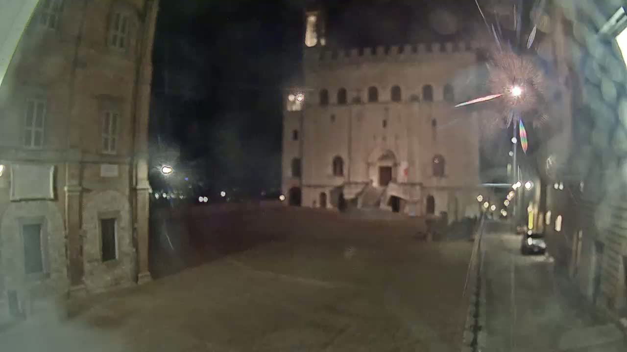 A nighttime view of a large, illuminated building across a mostly empty square, seen from a slightly elevated perspective down a street at night.