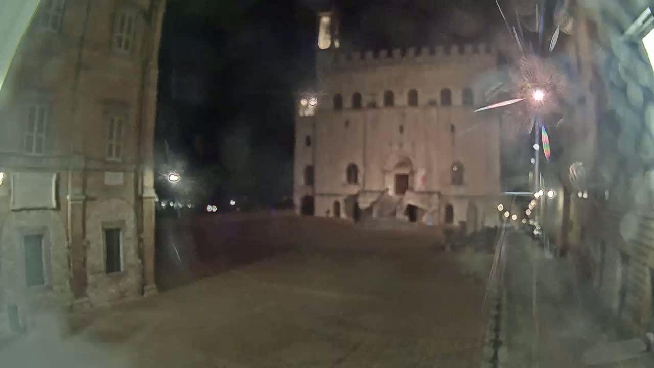 A nighttime view of a large, stone building with a staircase at its entrance, flanked by smaller buildings, in a mostly empty square.