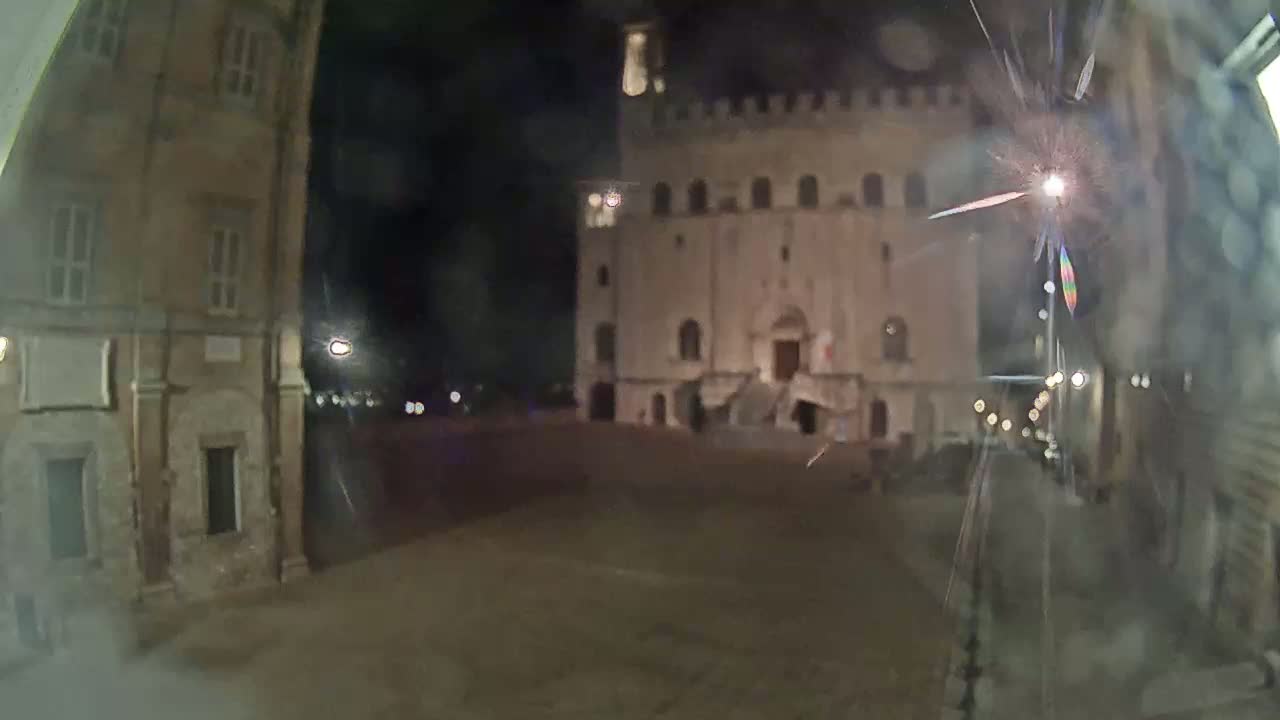 A nighttime view of a large, pale stone building with a staircase in a town square, seen through a slightly blurry, possibly rain-streaked, window.