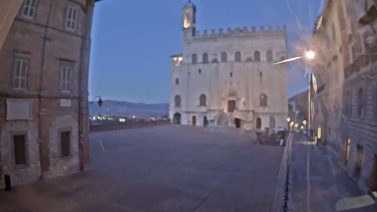 A large, light-colored stone building stands in a dimly lit town square at dusk, flanked by older, brick buildings.