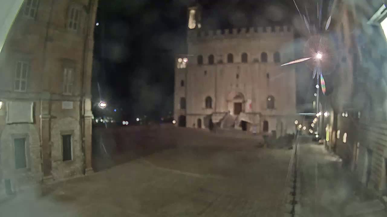 A nighttime view of a town square with an illuminated stone building, flanked by older buildings, under a dark sky.