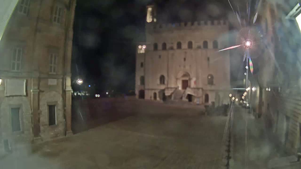 A nighttime view of a town square, featuring a large stone building with a staircase, seen through a slightly damaged window.