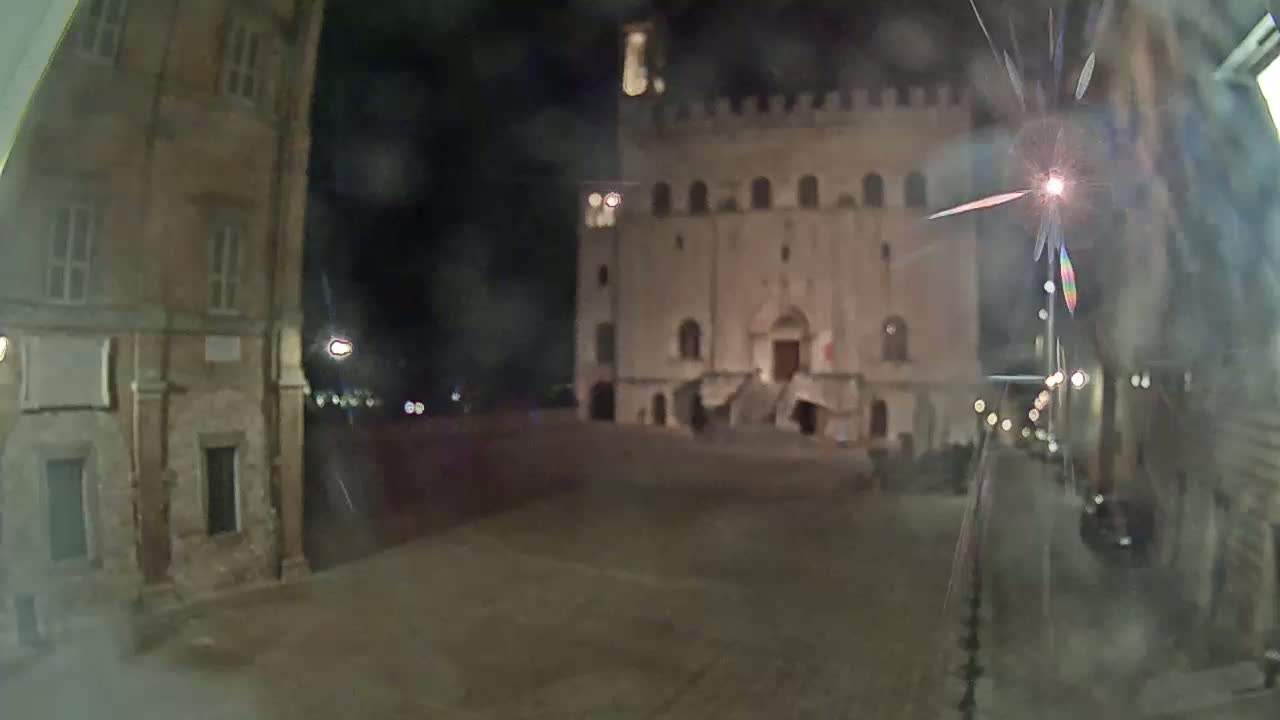 A large, empty square at night is flanked by old stone buildings, one of which has a prominent staircase leading to its entrance.