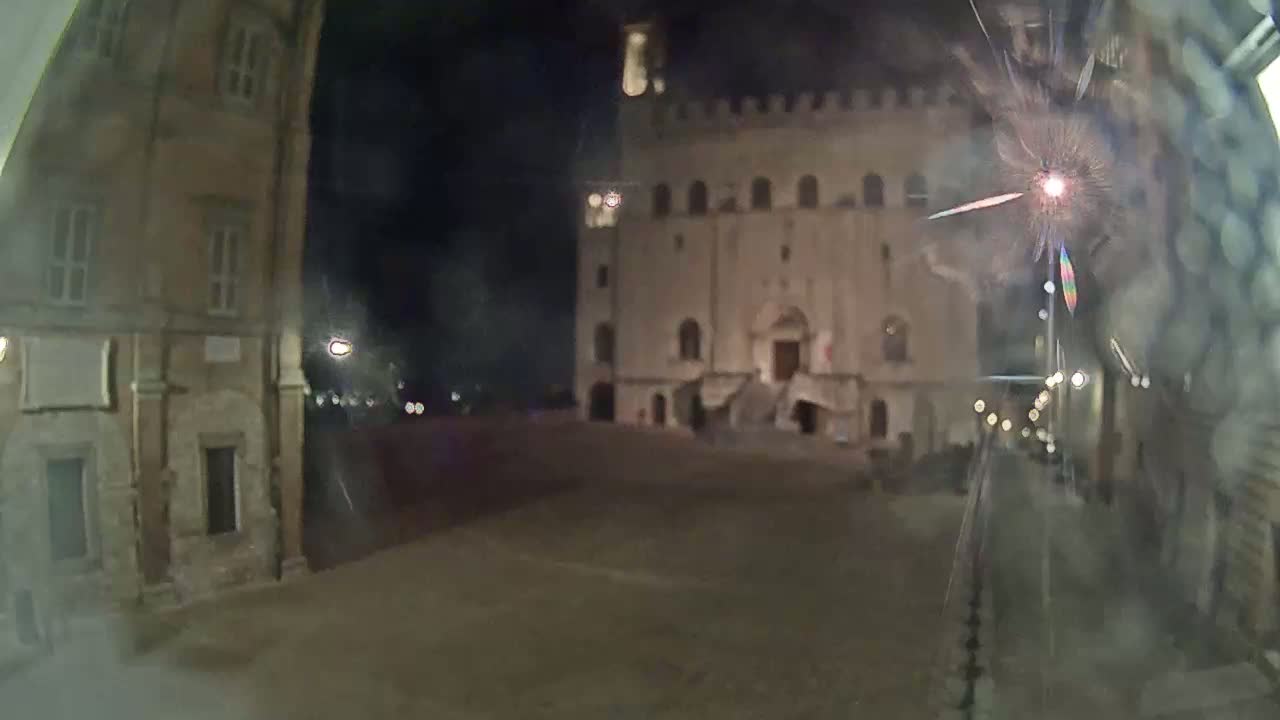 A nighttime view of a town square, featuring a large, light-colored building with steps, flanked by other buildings under a dark sky.