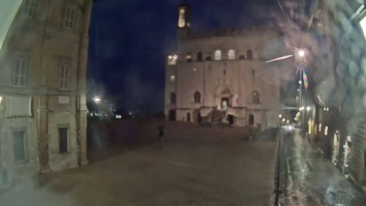 A large, light-colored building stands in a rain-slicked square at night, viewed from a perspective that seems to be through a rain-streaked window.