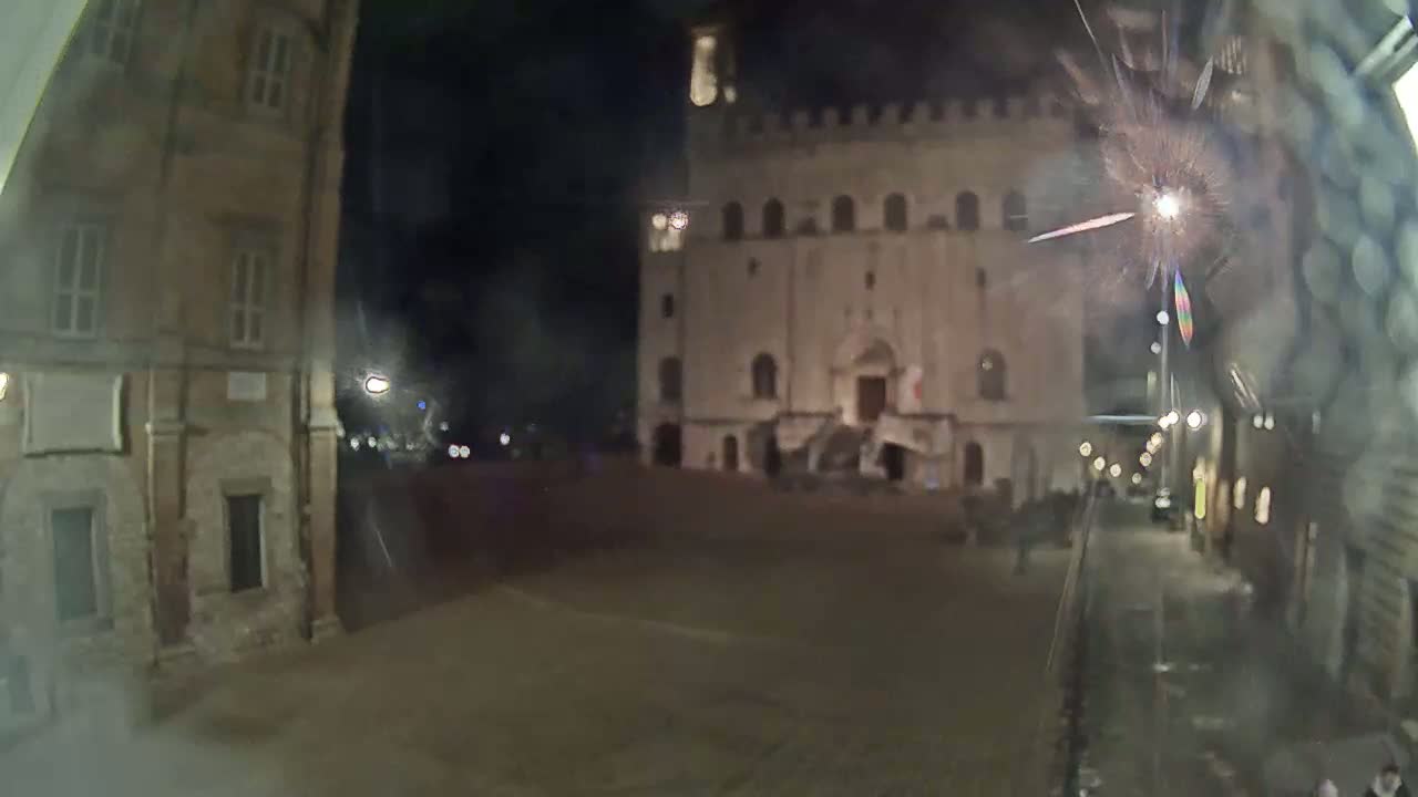A nighttime view of a town square with a large, illuminated building and fireworks in the distance, viewed from a slightly obscured perspective.