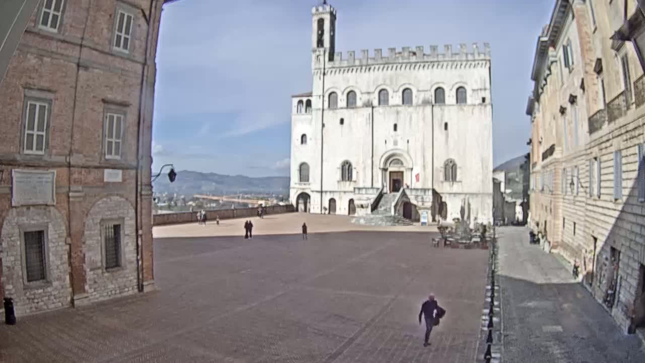 A large, mostly empty plaza is situated between two brick buildings, one of which is a tall, white stone structure with a staircase leading to its entrance, under a bright, sunny sky.