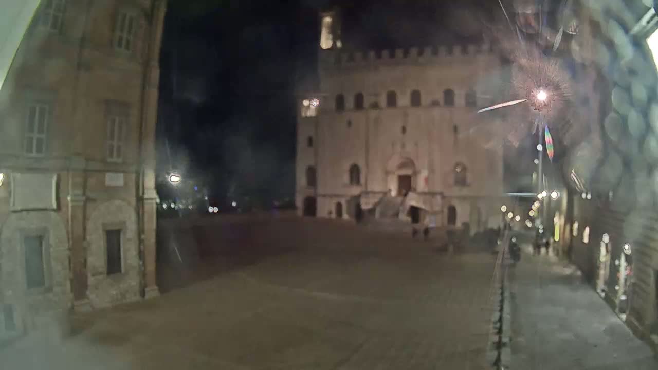 A nighttime view of a town square shows a large, light-colored building with stairs leading to its entrance, flanked by other buildings, under a dark, clear sky.