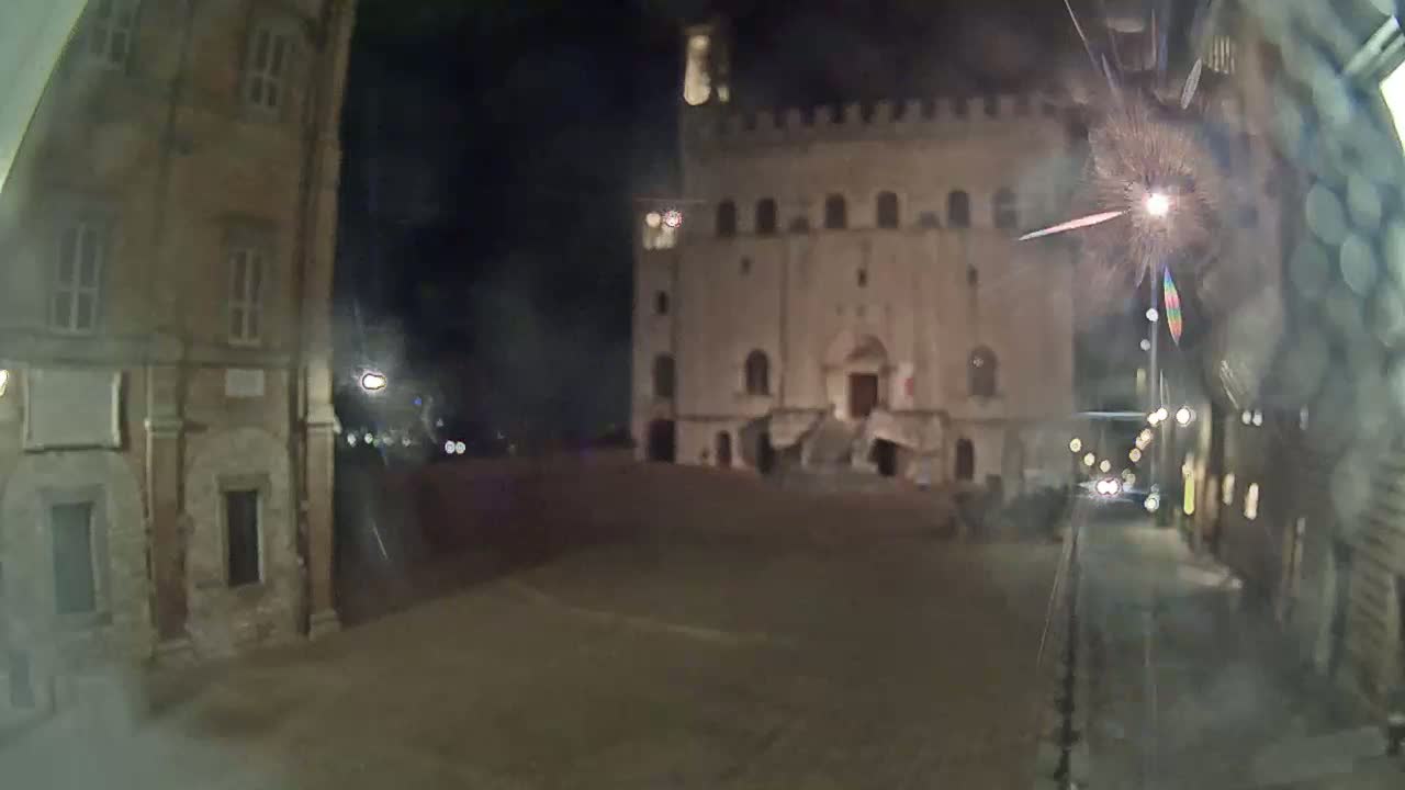 A nighttime view of a town square, featuring a large stone building with a staircase, flanked by other buildings, under dark, clear skies.