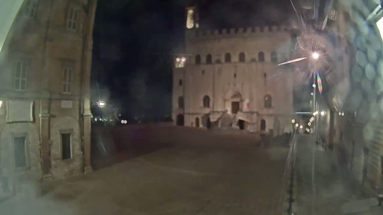 A large, illuminated stone building stands across a mostly empty square at night, with a blurry firework visible in the foreground.