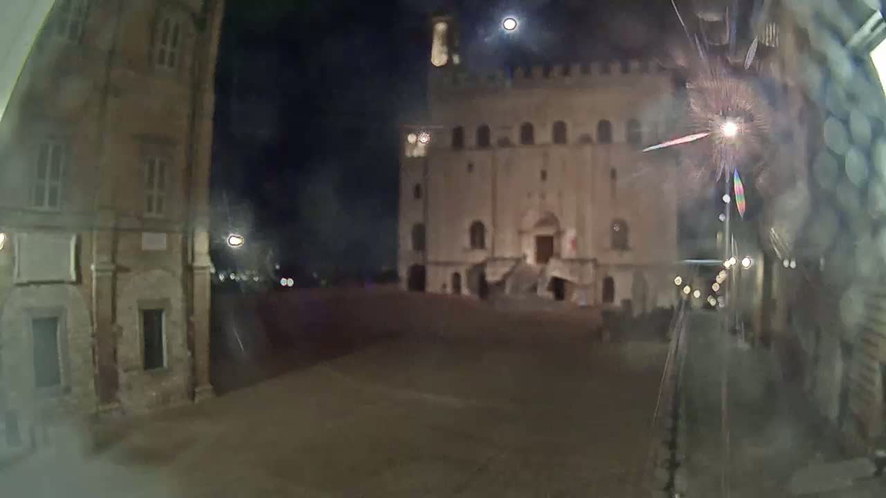 A large, light-colored building with a staircase stands across a mostly empty square at night, under a mostly clear sky with a visible moon.