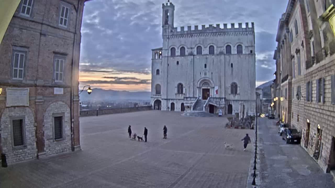 A large, light-colored stone building with a tower stands in a town square at sunrise, with several people and dogs in the square and cars parked along a street to the right.