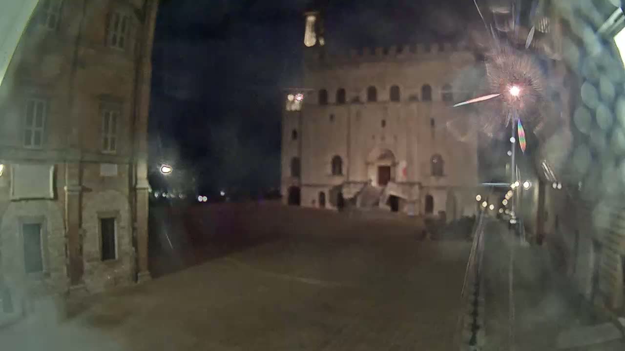 A nighttime view of a large, light-colored building with steps in front, situated in a square between two older, darker buildings, seen through a rain-streaked window.
