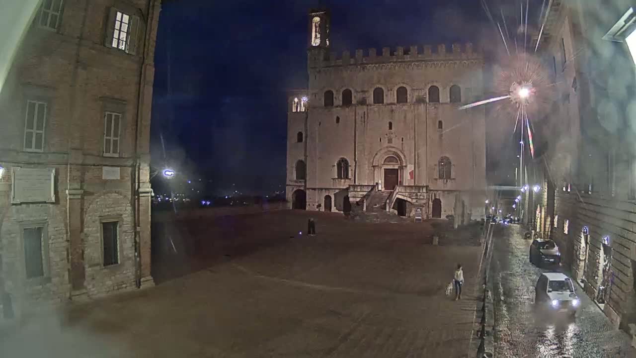 A large stone building with a staircase stands in a plaza at night under a dark, possibly rainy sky, with a few people and cars visible in the streets nearby.
