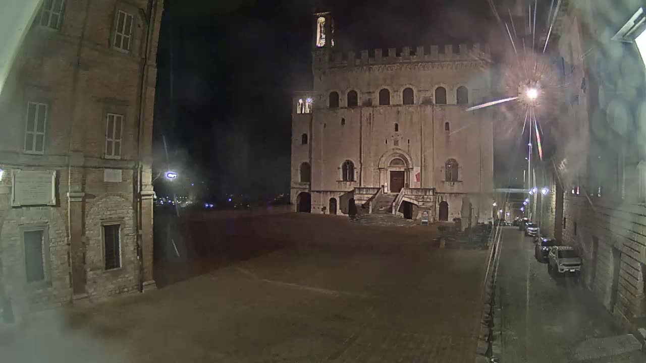 A large, stone building with a staircase stands in a dimly lit square at night, with a few cars visible on a nearby street.