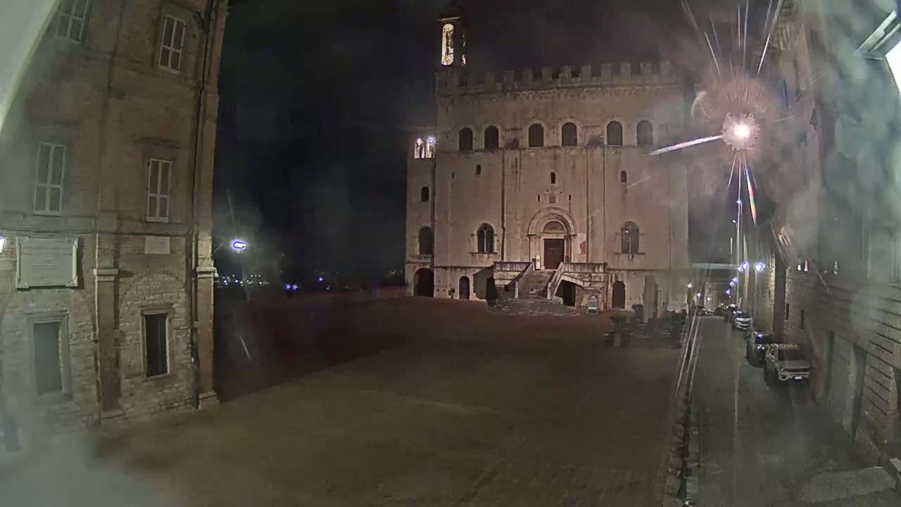 A large, stone building with a staircase is visible at night, with a street and other buildings also visible in the distance, under misty or smoky conditions.