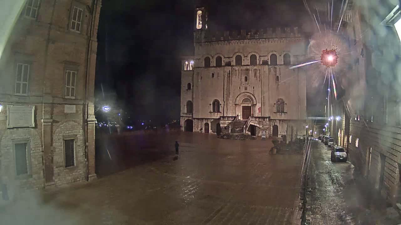 A large, flood-covered town square at night is dominated by a stone building with a staircase, flanked by other buildings under rainy conditions.