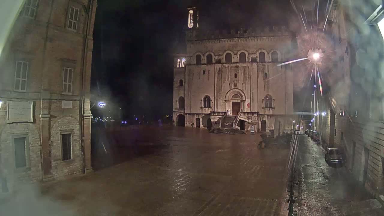 A large, stone building stands in a floodlit town square at night during a rainstorm, with water covering the cobblestone ground.