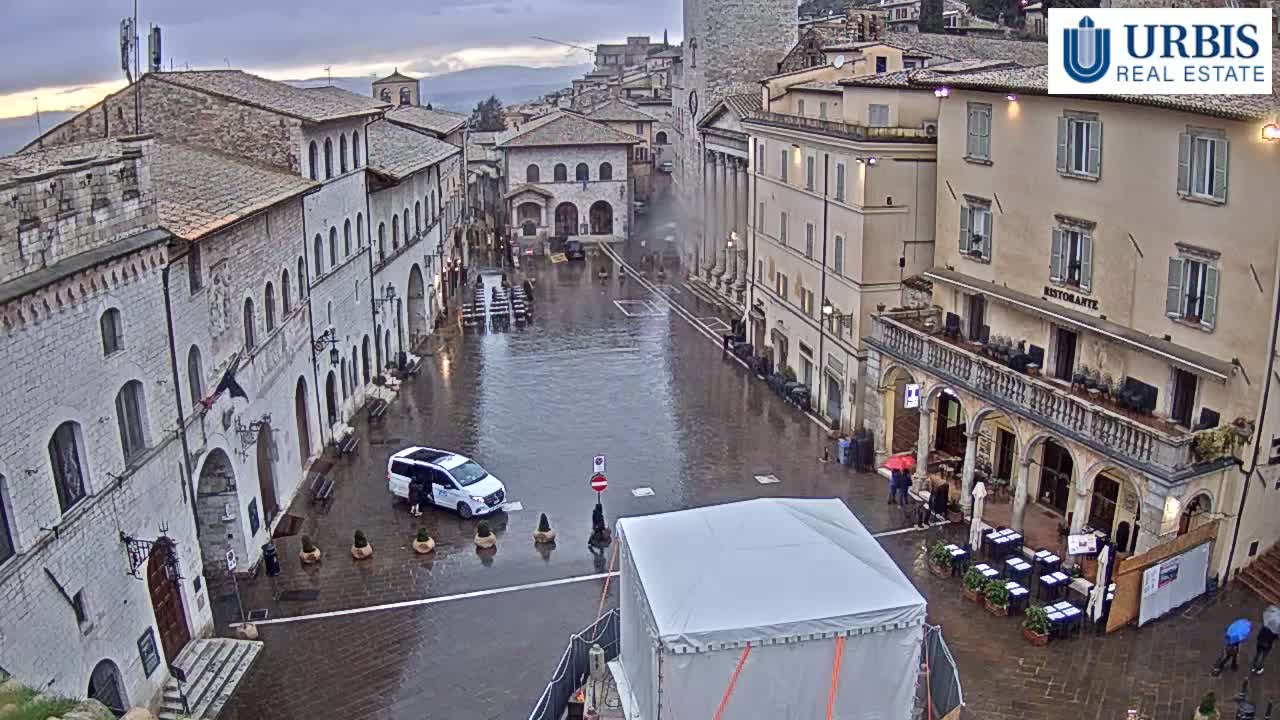 A rain-soaked town square in Italy is surrounded by historic stone buildings, with a single vehicle parked in the flooded plaza.