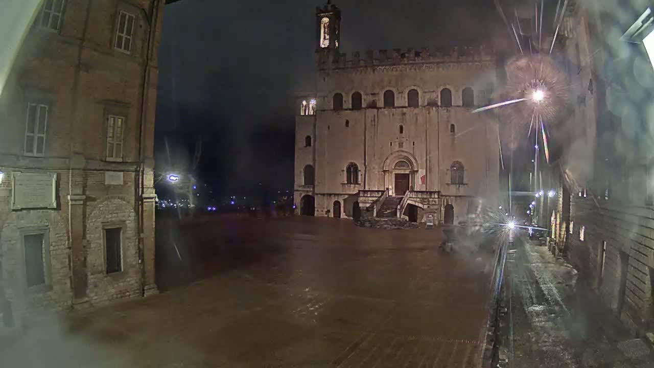 A large, stone building with a tower is situated in a town square at night during what appears to be a fireworks display, with wet pavement indicating recent rain or snow.
