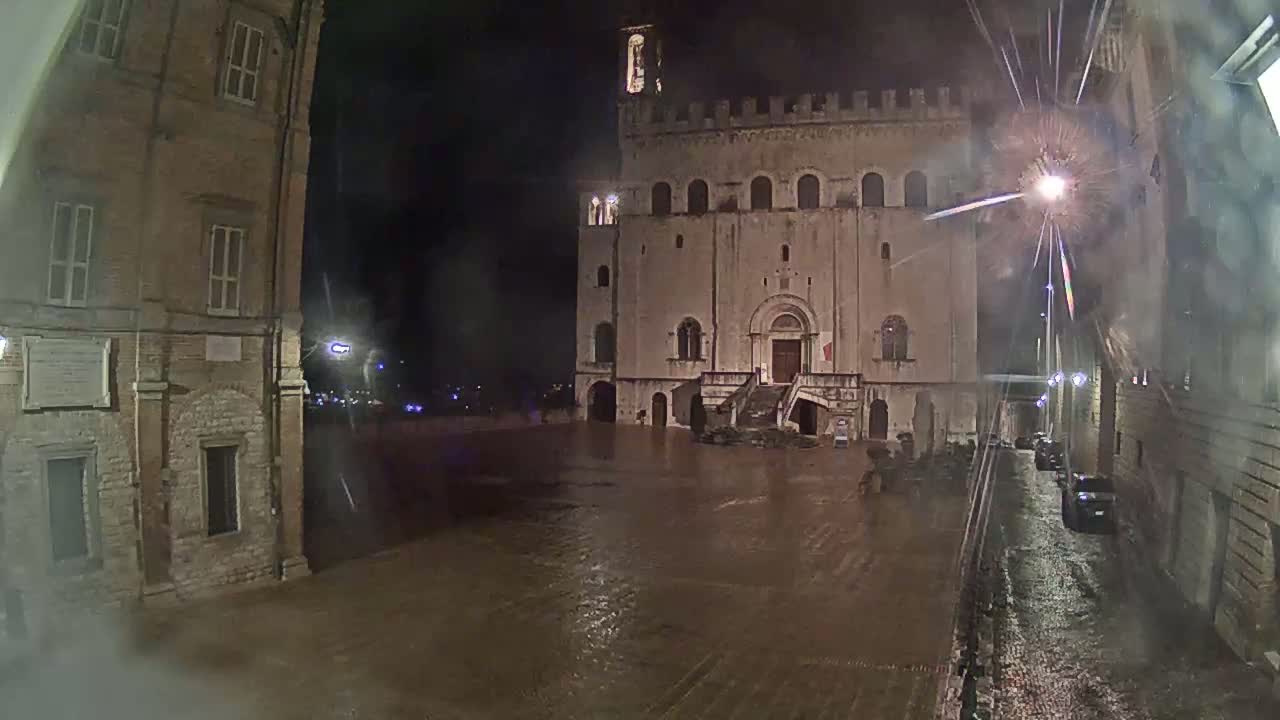 A large, flood-covered town square at night is flanked by old stone buildings under heavy rain.