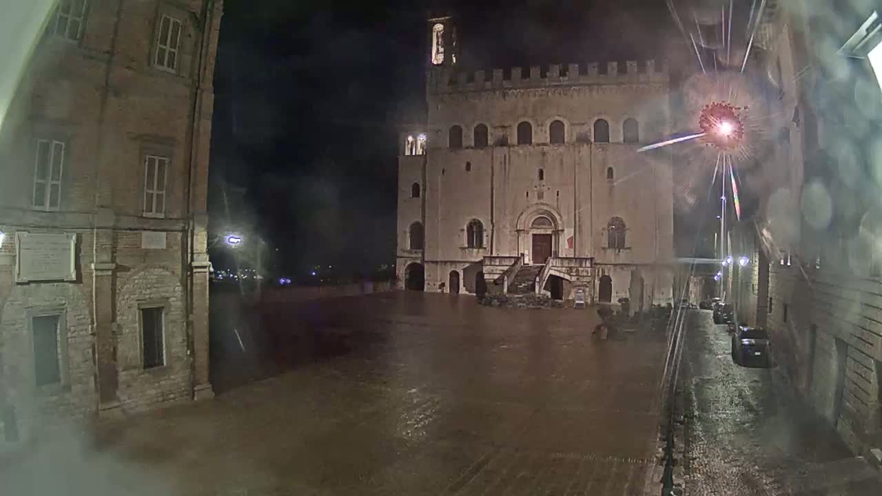 A large, stone building stands in a rain-soaked town square at night, with a street lined with buildings visible on the right.