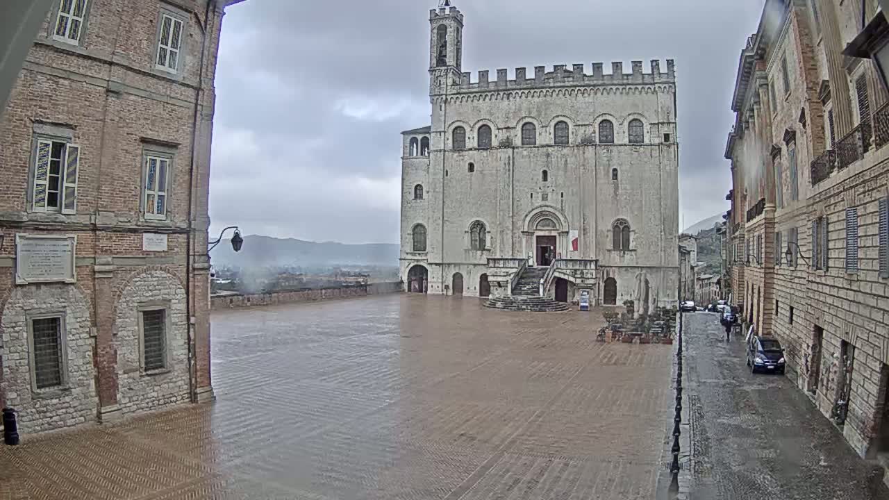 A large, stone palace sits in a town square under an overcast sky, with rain-slicked cobblestone streets and a wet plaza surrounding it.