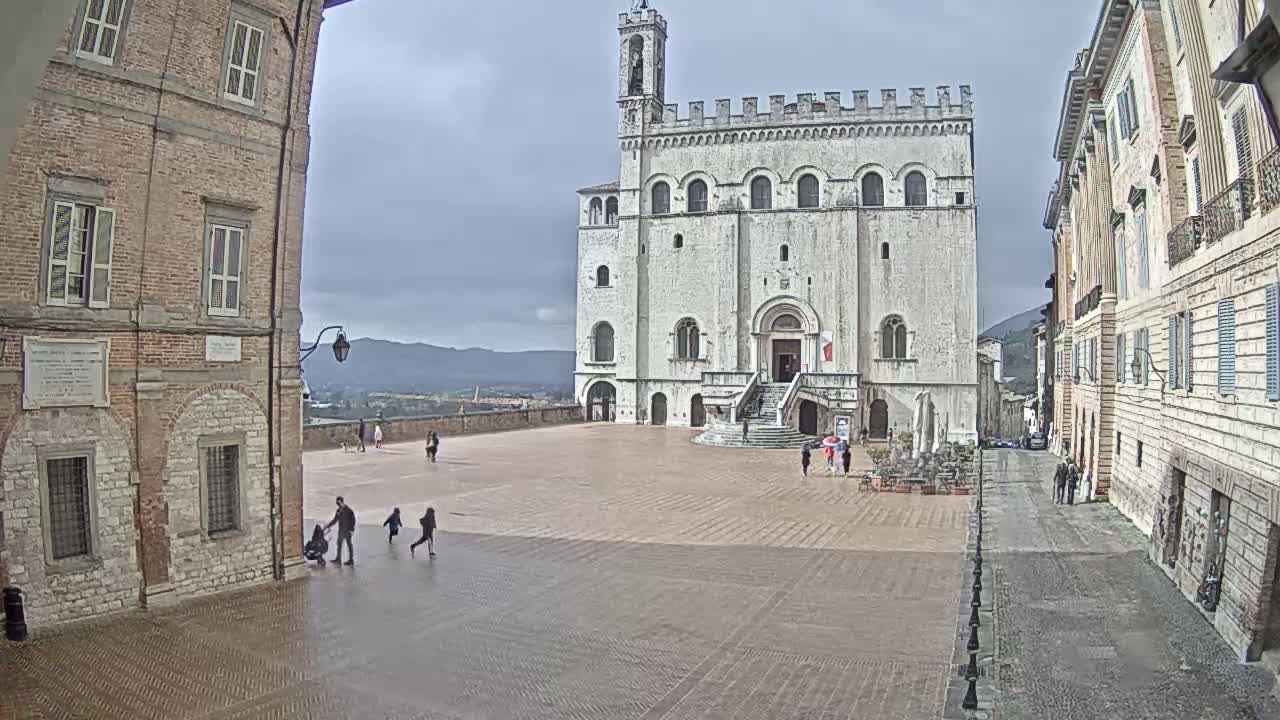 A large, white stone building stands in a town square on an overcast day, with a few people walking around and brick buildings lining the sides of the square.