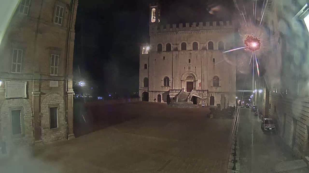 A large, stone building with a staircase stands in a dimly lit, empty square at night, with fireworks exploding in the background.