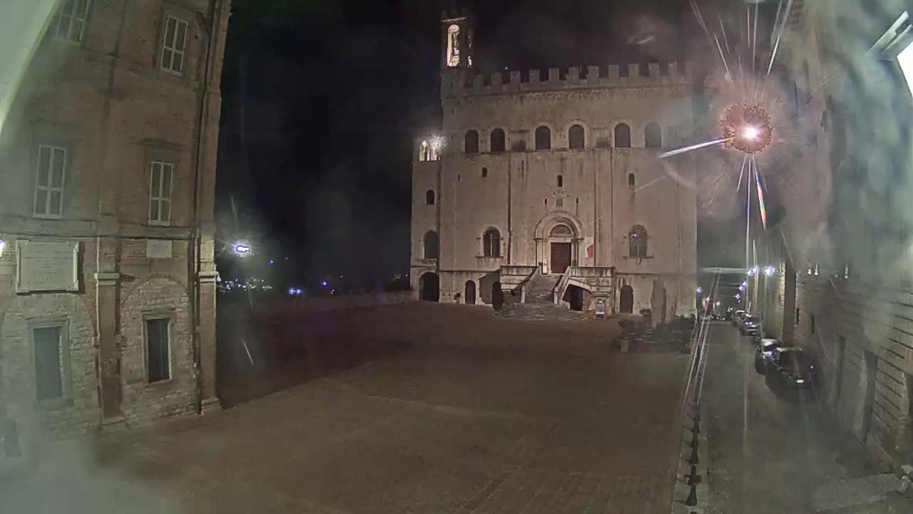 A large, stone building with a staircase and tower is visible in a dimly lit town square at night, with fireworks exploding in the background.