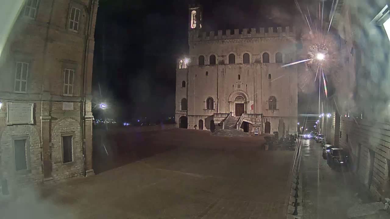 A large, stone building with a crenelated top stands in a square at night, illuminated by streetlights, with a few cars parked nearby.
