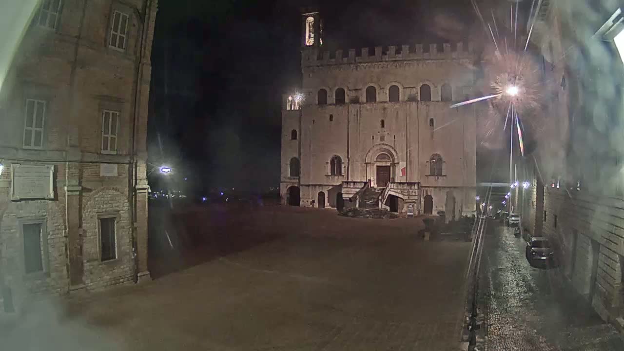 A large, stone building with a staircase stands in a square at night, with fireworks exploding in the sky and wet cobblestone streets visible.