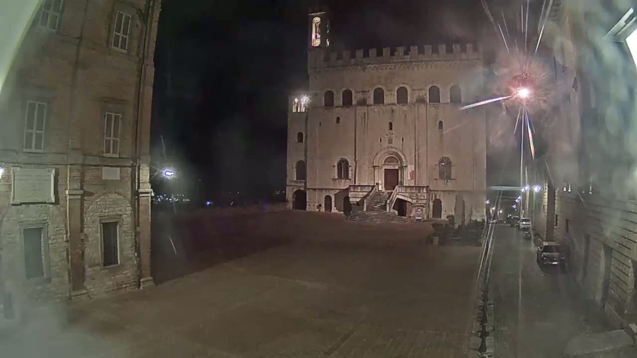 A large, stone building with a staircase and a clock tower is seen at night in a town square, with fireworks exploding in the sky.