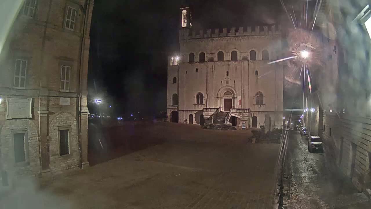 A large, stone building with a staircase stands in a dimly lit, empty square at night, under what appears to be rainy conditions.