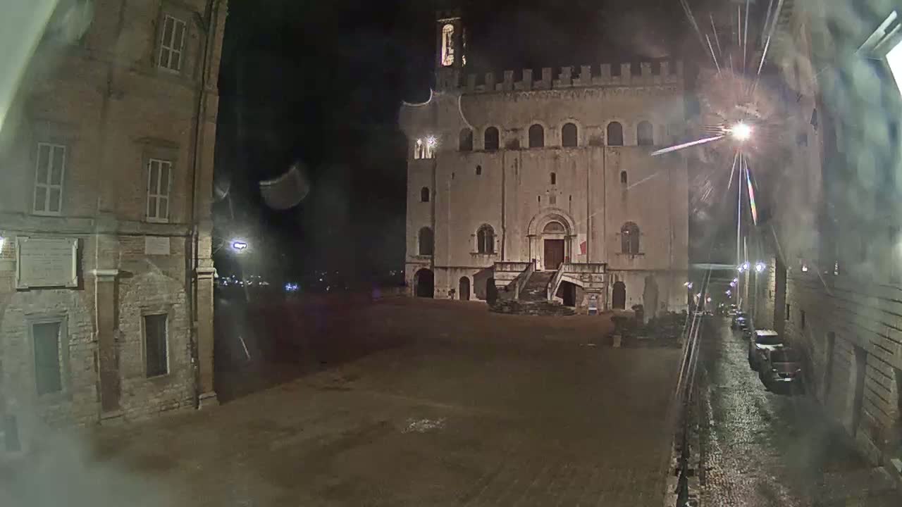 A large, stone building with a staircase stands in a dark, empty square at night, with rain-slicked streets visible in the foreground.