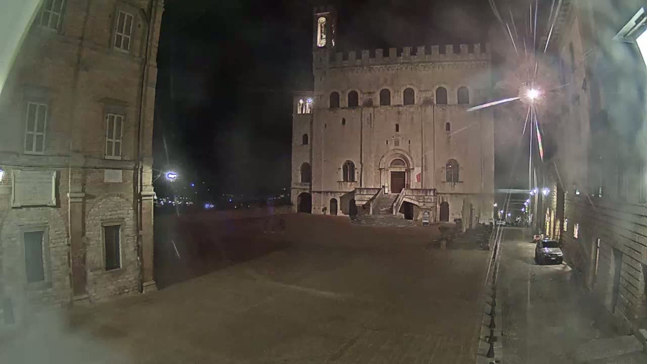 A large, stone building with a crenellated top is seen at night in a town square, with misty or hazy weather conditions.
