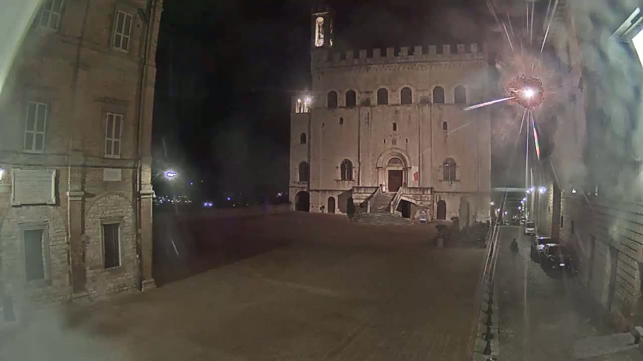A large, stone building with a staircase stands in a town square at night, with fireworks exploding in the sky.