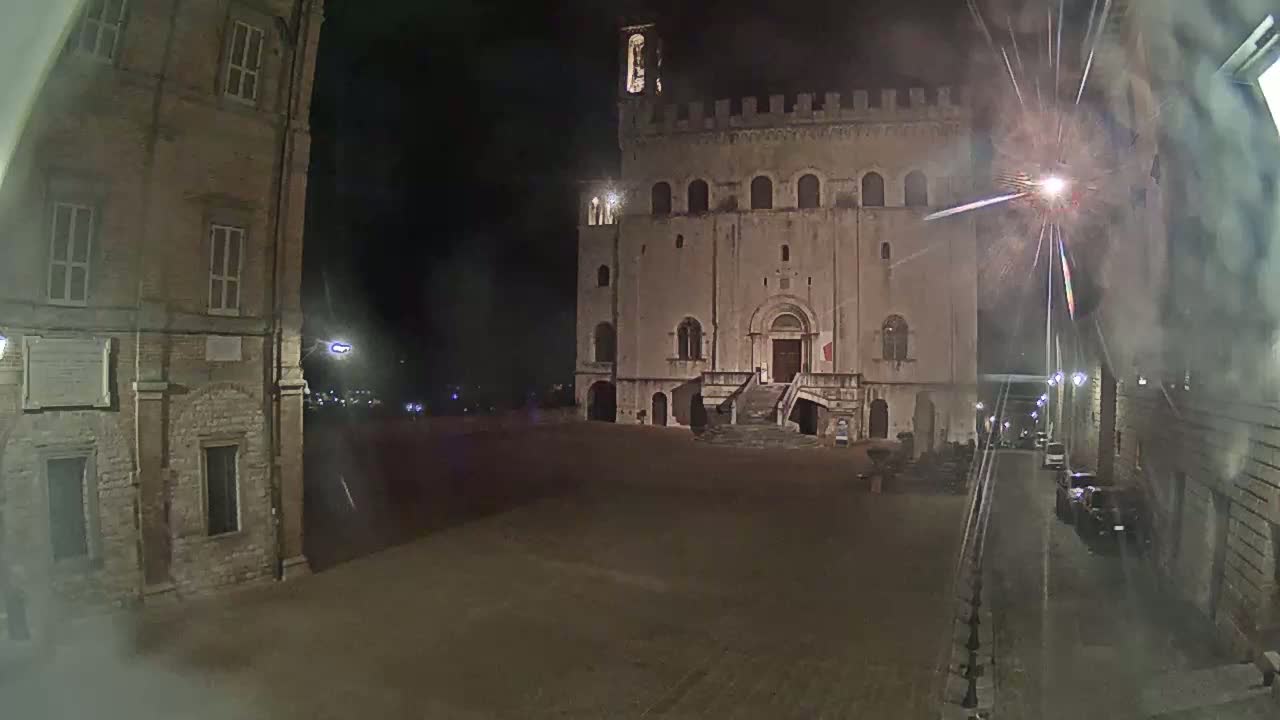 A large, stone building with a staircase stands in a dimly lit square at night, with fireworks visible in the sky.