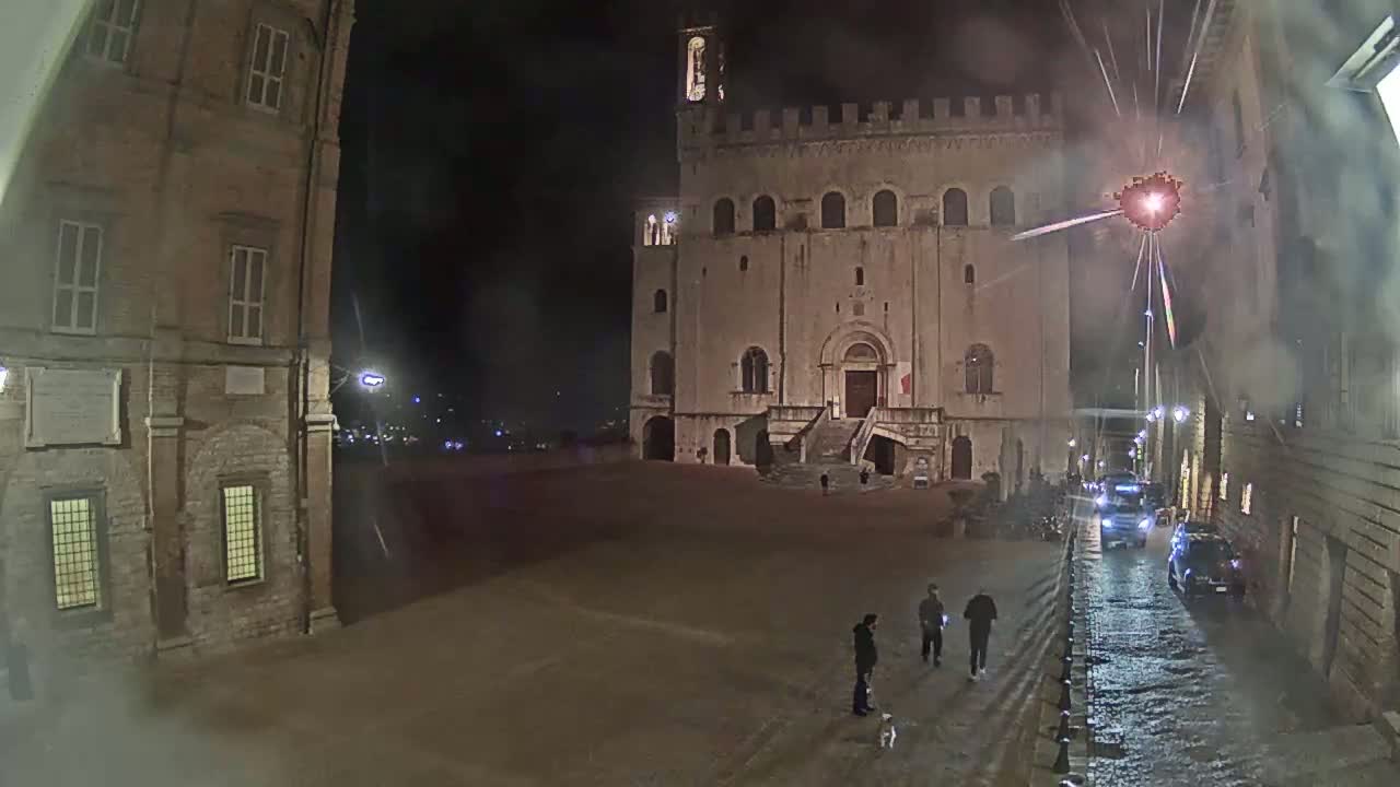 A large, stone building is visible in a nighttime square with a few people walking, and fireworks are exploding in the background under a dark sky.