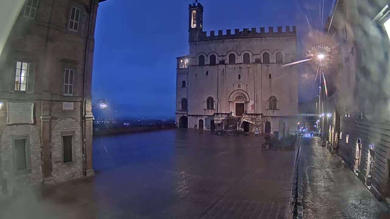 A large, light-colored stone building with a tower stands in a square at night during a light rain.