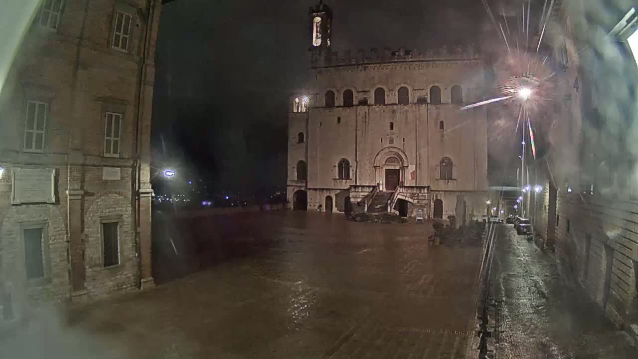 A large, stone building with a staircase stands in a rain-soaked square at night, flanked by other buildings under a dark, stormy sky.