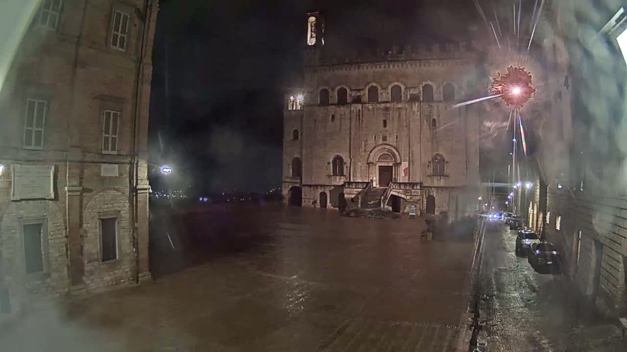 A large stone building stands in a rain-soaked square at night, with fireworks exploding in the background.