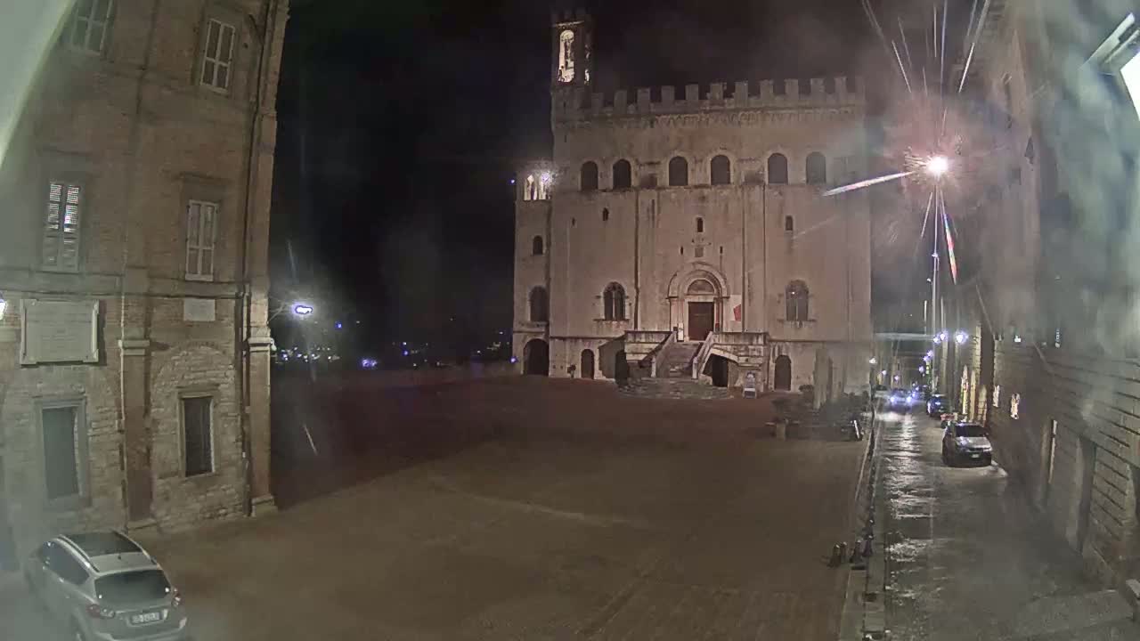 A large, stone building stands in a dimly lit, empty square at night, with a few cars visible on a nearby street under slightly wet conditions.