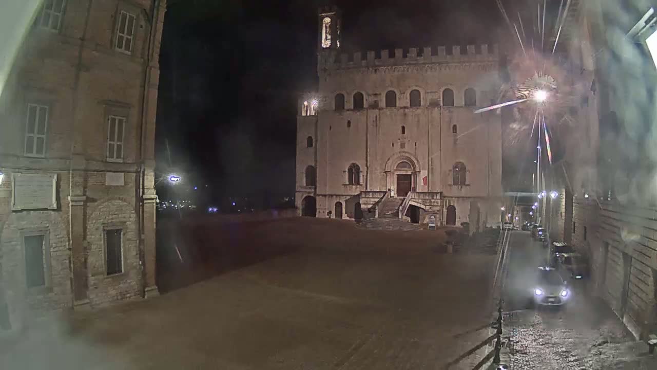 A large, stone building with a staircase stands in a mostly empty square at night, with fireworks exploding in the background and a wet street visible in the foreground.